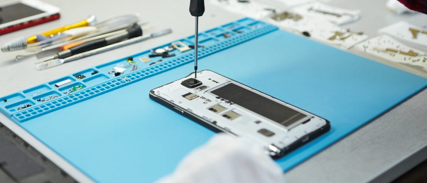Cropped shot of male technician in antistatic gloves using screwdriver to disassemble borken mobile phone, going to repair motherboard, sitting at his workplace in laboratory with needed equipment